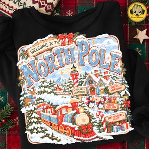 Puede incluir: Una sudadera negra con un gráfico colorido de una escena invernal del Polo Norte. El diseño incluye un tren rojo, un faro, pingüinos, un reno y el texto Welcome To The North Pole, Polar Lighthouse, Santa's Workshop, Penguin Parcel Crew y Free Delivery Depot.
