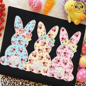 May include: Three bunny silhouettes with floral patterns in light blue, yellow, and pink. Each bunny has a decorative bow and a white, pearl-centered tail. The bunnies are on a black background, with Easter-themed decorations.