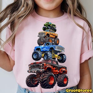 May include: Pink t-shirt featuring a stack of five colorful monster trucks. The trucks are in shades of red, blue, orange, green, and black, with large tires and detailed designs. The text "GrootStoree" is in the bottom right corner.