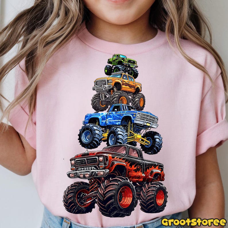 May include: Pink t-shirt featuring a stack of five colorful monster trucks. The trucks are in shades of red, blue, orange, green, and black, with large tires and detailed designs. The text "GrootStoree" is in the bottom right corner.