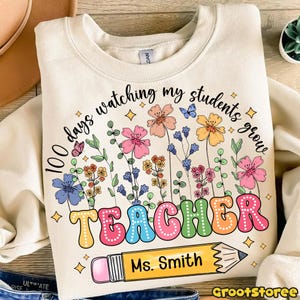 May include: Cream-colored sweatshirt with a colorful floral design and the text "100 days watching my students grow TEACHER Ms. Smith." The design includes butterflies, flowers, and a pencil.