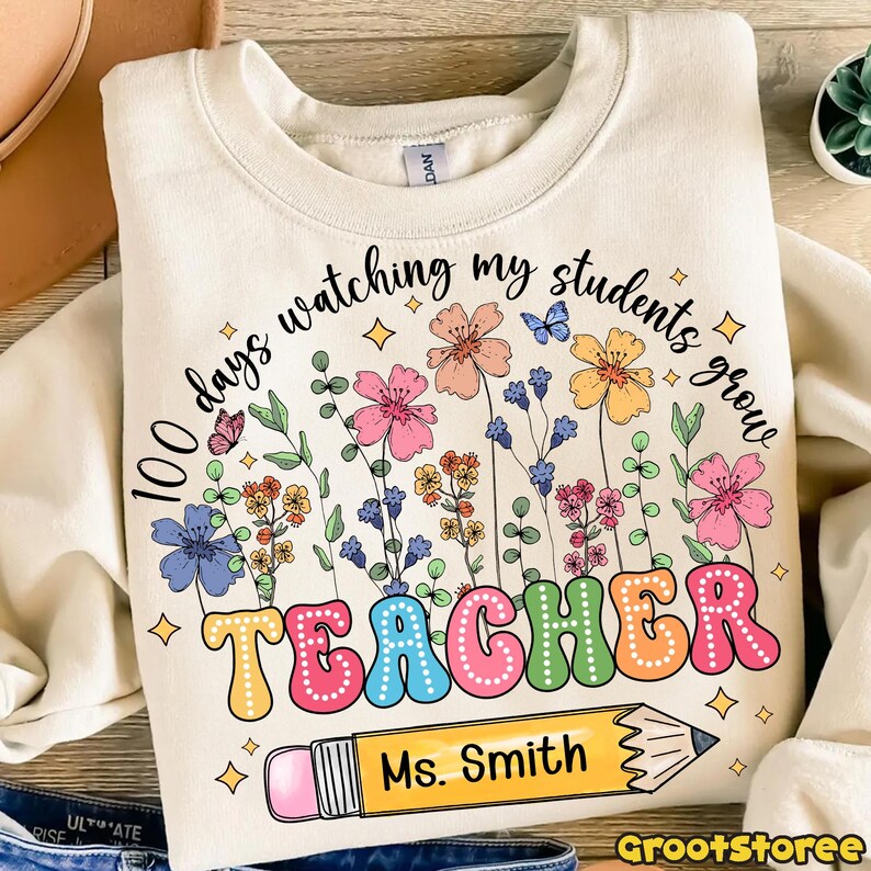 May include: Cream-colored sweatshirt with a colorful floral design and the text "100 days watching my students grow TEACHER Ms. Smith." The design includes butterflies, flowers, and a pencil.