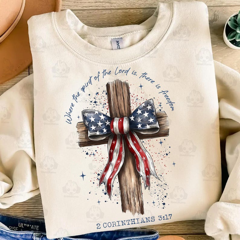 Może przedstawiać: Kremowa bluza z nadrukiem drewnianego krzyża z czerwoną, białą i niebieską kokardą. Tekst głosi: &bdquo;Where the spirit of the Lord is, there is freedom&rdquo; i &bdquo;2 Koryntian 3:17&rdquo;.