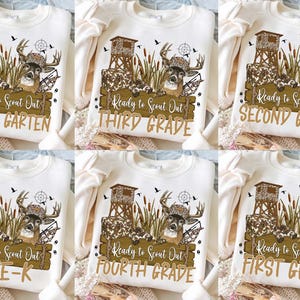 May include: A cream-colored sweatshirt featuring a graphic of a deer wearing a camouflage hat, a hunting stand, and cattails. The text reads Ready to Scout Out followed by various grade levels including Kindergarten, First Grade, Second Grade, Third Grade, Fourth Grade, and Pre-K.