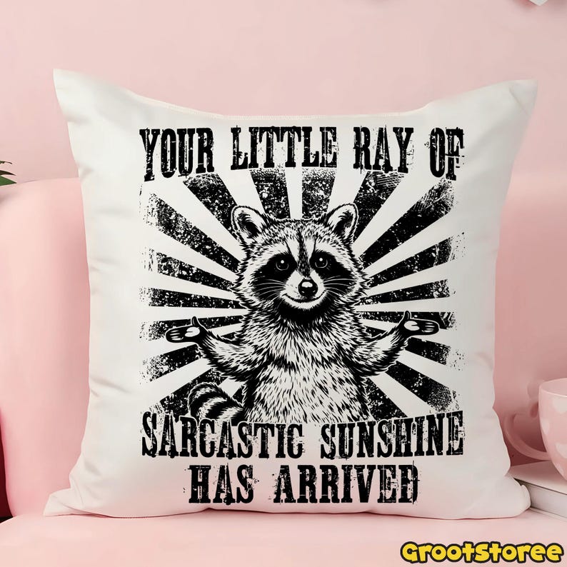 May include: White square cushion featuring a black and white illustration of a raccoon with its arms out. The text reads "YOUR LITTLE RAY OF SARCASTIC SUNSHINE HAS ARRIVED". The design has a distressed, vintage aesthetic.
