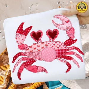 May include: A white tote bag featuring a patchwork crab design in shades of pink and red. The crab has heart-shaped eyes and various patterns, including polka dots, stars, and hearts. The design is whimsical and beach-themed.