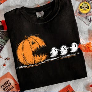 May include: Black t-shirt featuring a Halloween design. An orange jack-o'-lantern with a jagged mouth is on the left, with three white cartoon ghosts trailing behind it. The design is a simple line drawing style.