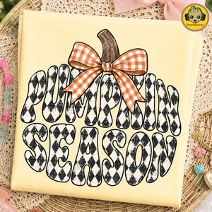 May include: A pale yellow t-shirt featuring a large graphic of a pumpkin shape filled with a black and white diamond pattern. The text PUMPKIN SEASON is written in a wavy font within the pumpkin, topped with an orange and white gingham bow.