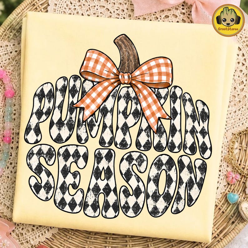 May include: A pale yellow t-shirt featuring a large graphic of a pumpkin shape filled with a black and white diamond pattern. The text PUMPKIN SEASON is written in a wavy font within the pumpkin, topped with an orange and white gingham bow.