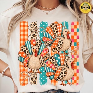 May include: A cream-colored t-shirt featuring a graphic of three turkeys with textured, multi-colored feathers in patterns of leopard print, orange plaid, and teal polka dots, set against vertical stripes of orange and teal checkered squares.