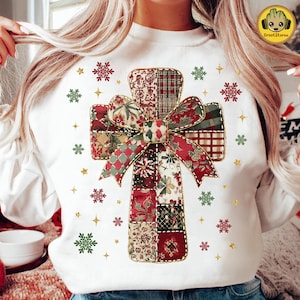 May include: White sweatshirt featuring a patchwork cross design with a large bow. The cross is made of various patterned fabrics in red, green, and cream. The design is surrounded by red, green, and gold snowflake and star accents.