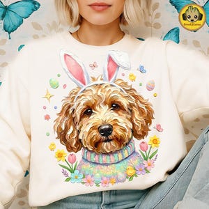 May include: Cream-colored sweatshirt featuring a dog wearing bunny ears and a pastel-colored sweater. The dog is surrounded by Easter-themed graphics, including eggs, flowers, and butterflies. The sweatshirt has a crew neck and long sleeves.
