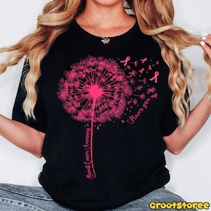 May include: Black t-shirt featuring a pink dandelion design with the words "Breast Cancer Awareness" and "Never give up." Pink ribbons are scattered around the dandelion. The shirt is a crew neck.
