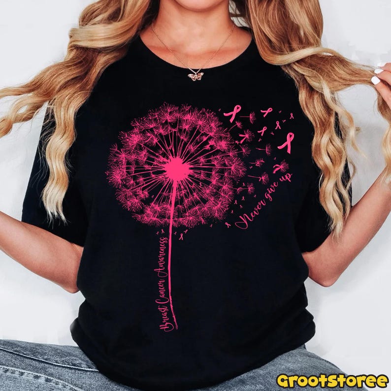 May include: Black t-shirt featuring a pink dandelion design with the words "Breast Cancer Awareness" and "Never give up." Pink ribbons are scattered around the dandelion. The shirt is a crew neck.