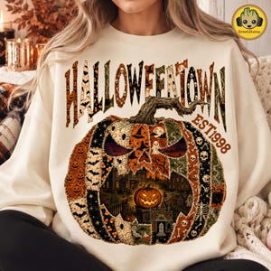 May include: A cream-colored crewneck sweatshirt featuring a large graphic print. The text reads HALLOWEENTOWN EST 1998. Below, a large pumpkin illustration is composed of various Halloween-themed patterns, including bats, ghosts, and skulls.