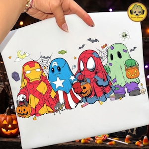 Op de afbeelding: Een wit t-shirt met een grafische print van vier personages verkleed als spoken voor Halloween. De figuren lijken op Iron Man, Captain America, Spider-Man en een groen spook, die elk een pompoen-snoepemmer vasthouden.