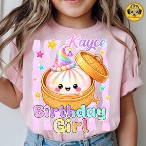 May include: A light pink t-shirt featuring a graphic of a smiling, glittery bao bun inside a bamboo steamer basket. The bun wears a colourful party hat. The text reads Kayce Birthday Girl in a gradient font, surrounded by small yellow, pink, and blue stars.