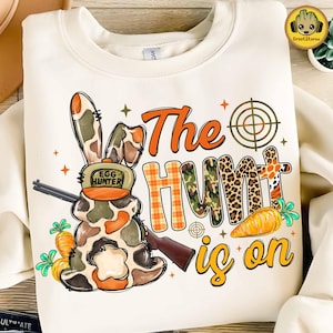 May include: Cream-colored sweatshirt with a graphic featuring a bunny in camouflage, a rifle, and the text "The Hunt is on." The word "Hunt" is designed with camouflage, leopard print, and plaid patterns. Carrots and a target are also included.