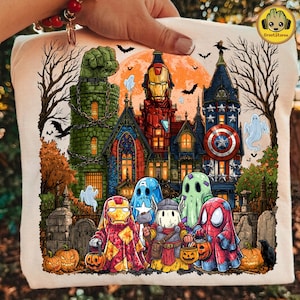 May include: A cream-colored t-shirt featuring a graphic print of a haunted mansion with superhero-themed architecture and characters dressed in ghost costumes, including Iron Man, Thor, and Spider-Man, set against a large orange moon.