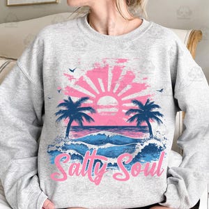 May include: A gray sweatshirt with a pink and blue beach scene graphic. The graphic features palm trees, a sunset, and waves. The text "Salty Soul" is written in pink below the graphic.