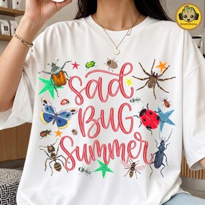 May include: White t-shirt with a colourful graphic design featuring various insects and the text "Sad Bug Summer" in red cursive. The design includes a butterfly, ladybug, and other bugs.