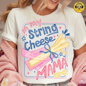 May include: White t-shirt with a colorful graphic that reads "In my String Cheese Mama Era." The design features a bundle of string cheese tied with a blue ribbon, and a pink and blue border.