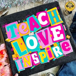 May include: Dark gray t-shirt with the words "TEACH LOVE INSPIRE" in large, colorful, block letters. The letters are outlined in teal and white, with pink, yellow, and white fill. The design includes school-themed icons.