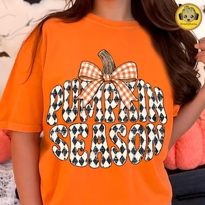 May include: An orange short-sleeved t-shirt featuring a large graphic of a pumpkin shape filled with a black and white diamond pattern. The text PUMPKIN SEASON is written in bold letters within the pumpkin, topped with an orange and white gingham bow.