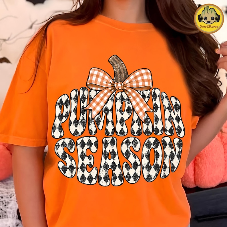 May include: An orange short-sleeved t-shirt featuring a large graphic of a pumpkin shape filled with a black and white diamond pattern. The text PUMPKIN SEASON is written in bold letters within the pumpkin, topped with an orange and white gingham bow.