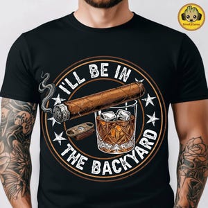 May include: A black t-shirt featuring a circular graphic with a brown cigar, a glass of whiskey with ice cubes, and a cigar cutter. The text "I'LL BE IN THE BACKYARD" is printed in white around the central illustration, which is accented by white stars.