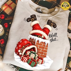 May include: A light beige sweatshirt featuring a graphic of Santa Claus stuck upside down in a brick chimney, with legs in red pants and black boots visible. A red gift sack and several wrapped presents sit below, with the text Well, shit...