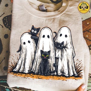 May include: Beige sweatshirt featuring a Halloween design with three white ghost figures and three black cats. The cats have yellow eyes and are positioned around the ghosts. The design is printed on a folded sweatshirt.