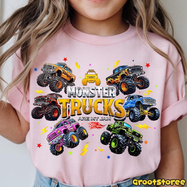 May include: Pink t-shirt featuring a colorful graphic of monster trucks. The design includes various monster trucks in orange, black, blue, purple, and green, with the text "MONSTER TRUCKS ARE MY JAM". Lightning bolt and star accents complete the design.