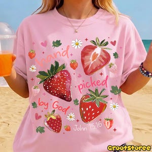 May include: Pale pink t-shirt with a strawberry design. The shirt features the text "hand picked by God" and "John 15:16" with illustrations of strawberries, daisies, and hearts. The shirt is a light pink colour.