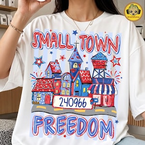 May include: White t-shirt featuring a colorful graphic of a small town with houses, a water tower, and the text "SMALL TOWN FREEDOM" in red and blue. The graphic includes the number "240966".