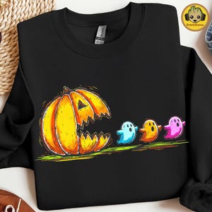 Puede incluir: Sudadera negra con un diseño de Halloween. A la izquierda, una gran calabaza naranja con boca dentada, seguida de cuatro fantasmas de colores. Los fantasmas son azules, amarillos, naranjas y rosas.