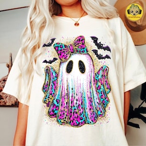 May include: Cream-colored t-shirt featuring a Halloween ghost design. The ghost has a bow and accents in pink, blue, and gold leopard print. Black bats and gold glitter surround the ghost, creating a festive look.