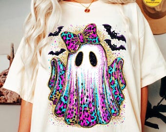 May include: Cream-colored t-shirt featuring a Halloween ghost design. The ghost has a bow and accents in pink, blue, and gold leopard print. Black bats and gold glitter surround the ghost, creating a festive look.