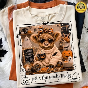 May include: A cream-coloured t-shirt featuring a graphic of a fluffy highland cow wearing heart-shaped sunglasses and a patterned bow. The cow holds a jack-o'-lantern mug. Text reads "STAY COZY AND SPOOKY" and "just a few spooky things".