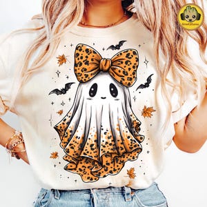 May include: A cream-colored t-shirt featuring a cartoon ghost with a leopard print bow and trim. The ghost has a friendly face and is surrounded by bats, stars, and autumn leaves. The shirt has the GrootStore logo.
