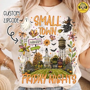 May include: A cream-colored t-shirt featuring a graphic print with the text SMALL TOWN FRIDAY NIGHTS, a football field, a water tower, ghosts, bats, flowers, and a sign with the number 686889. The design includes orange and purple accents.