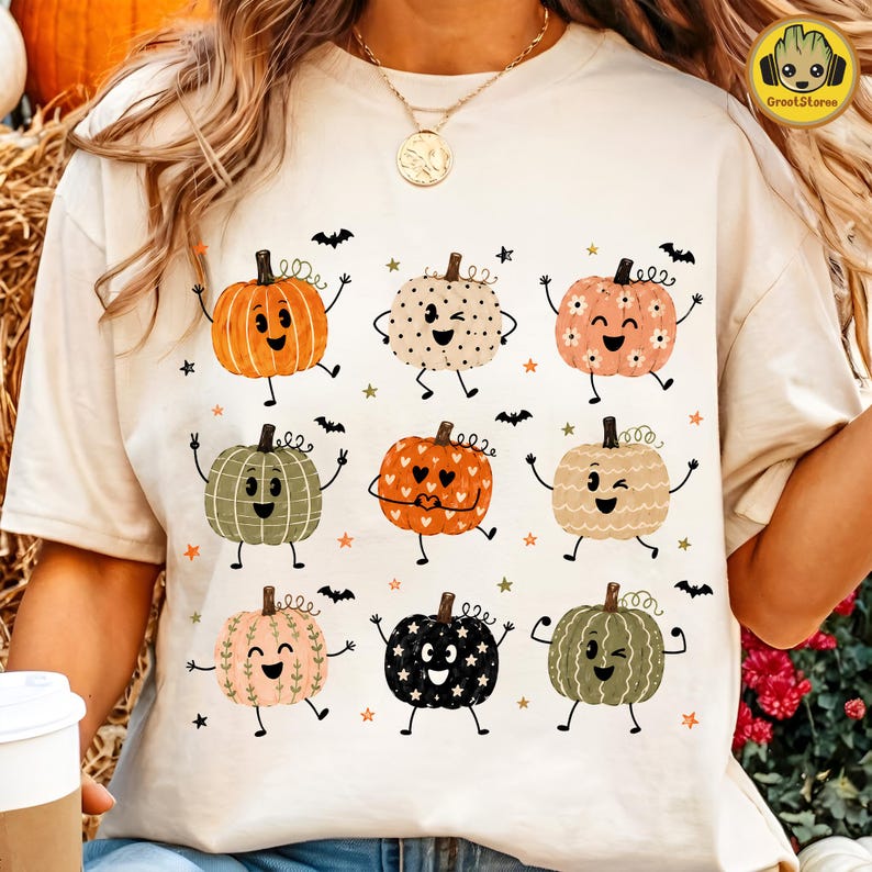May include: A cream-colored t-shirt featuring a graphic print of nine stylized pumpkins with faces, arms, and legs. The pumpkins are decorated with various patterns including hearts, stars, and flowers, interspersed with small black bats and stars.