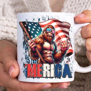 May include: White ceramic mug featuring a patriotic design. The mug displays a muscular Bigfoot character wearing sunglasses with an American flag design, holding a flag, and flashing a rock-on hand gesture. The word 'MERICA' is printed in red, white, and blue.