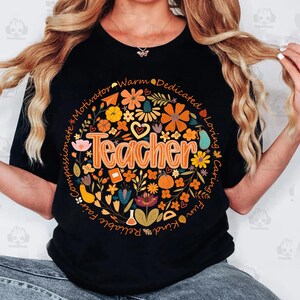 May include: Black t-shirt with a floral design in a circular pattern. The design includes the word "Teacher" in orange, surrounded by flowers, leaves, and words like "compassionate" and "loving".