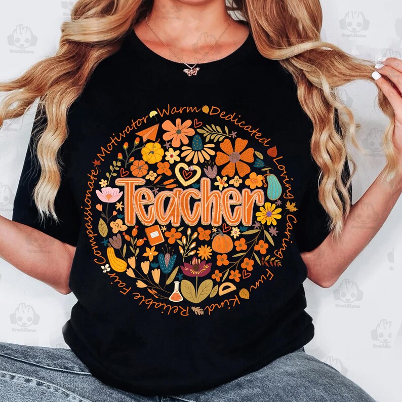May include: Black t-shirt with a floral design in a circular pattern. The design includes the word "Teacher" in orange, surrounded by flowers, leaves, and words like "compassionate" and "loving".
