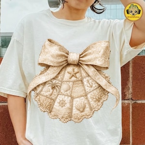 May include: Cream-colored t-shirt featuring a large, detailed seashell design with a bow. The seashell design incorporates nautical elements like a starfish, anchor, and palm tree. The shirt is a comfortable, casual style.