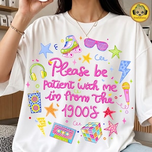 May include: White t-shirt with a colorful 1990s theme. The shirt features a graphic with the text "Please be patient with me, I'm from the 1900's" and illustrations of a roller skate, headphones, and a Rubik's Cube.