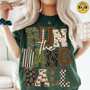 May include: A dark green t-shirt featuring the text RUN THE DAMN BALL in large block letters. Each letter has a unique pattern including football plays, helmets, stripes, mesh, footballs, pennants, and a checkerboard design in brown and green.