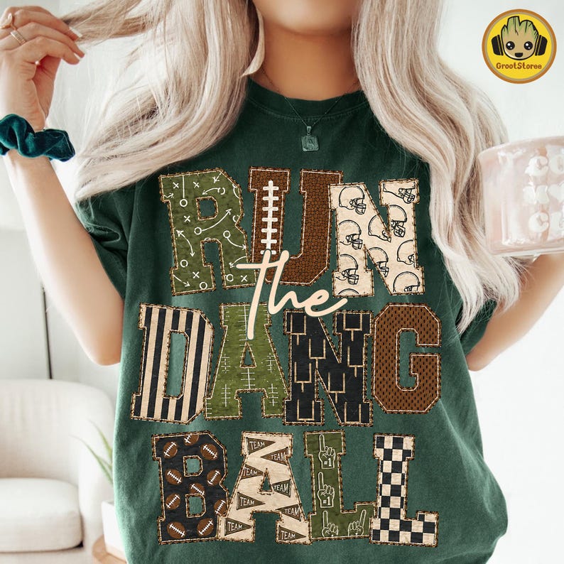 May include: A dark green t-shirt featuring the text RUN THE DAMN BALL in large block letters. Each letter has a unique pattern including football plays, helmets, stripes, mesh, footballs, pennants, and a checkerboard design in brown and green.