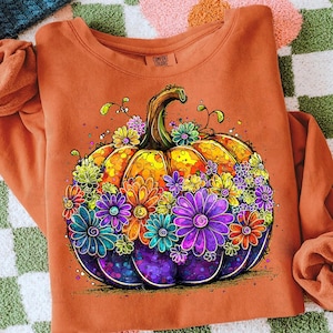 May include: An orange sweatshirt featuring a colorful, detailed illustration of a pumpkin decorated with vibrant flowers. The pumpkin has a yellow and orange body with a green stem, and is surrounded by purple, blue, and orange flowers.
